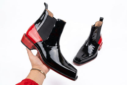 Black patent leather ankle boots with red soles held by a hand on a white background